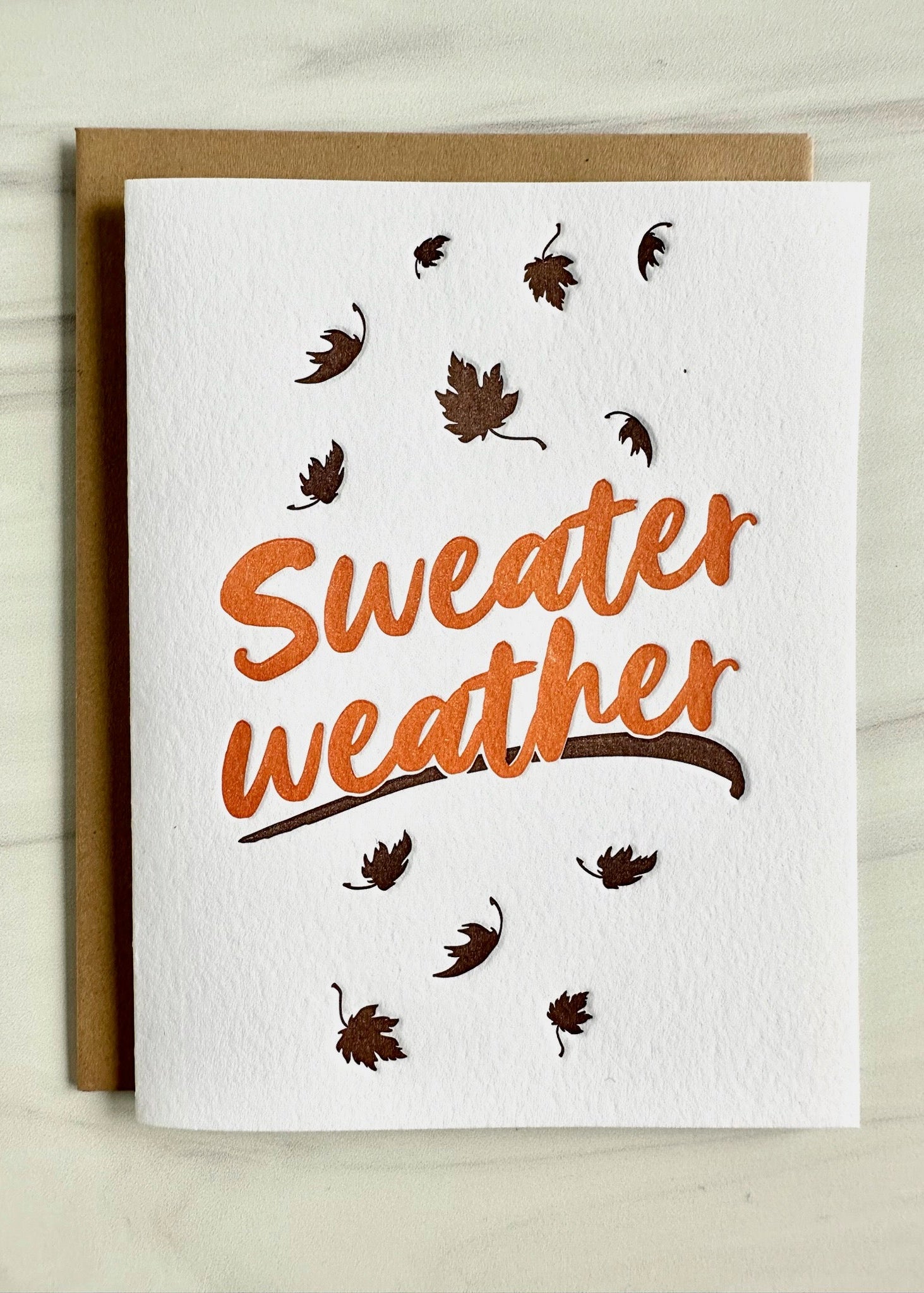 A greeting card with the phrase 'Sweater Weather' printed in orange on a white background, surrounded by brown illustrations of leaves.